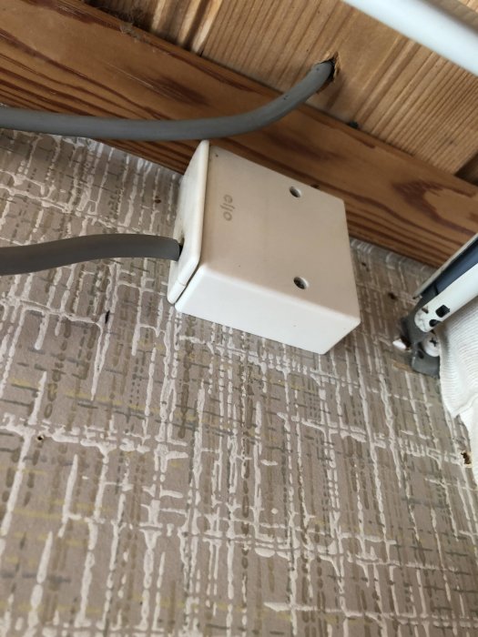 En vit, kvadratisk elektrisk dosa monterad på en vägg nära taket, med kablar anslutna, bakom en gardin.