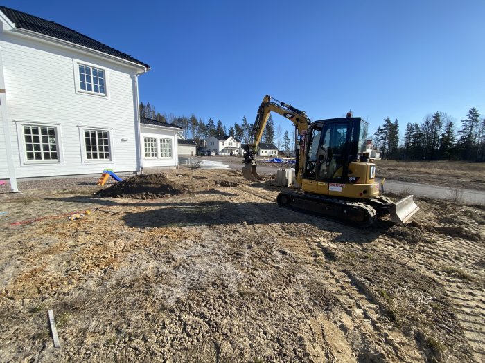 Grävmaskin bredvid nybyggd villa gräver för trädäck och pool i soligt, gräsbevuxet område med leksaker i förgrunden.