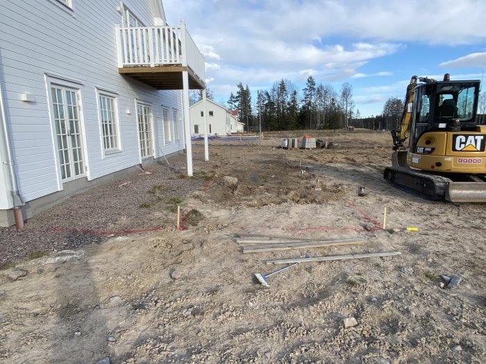 Uppgrävd utemiljö med en grävmaskin och märkstolpar för plintar vid hus.