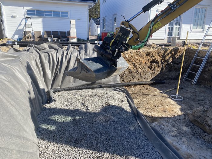 Grävmaskin som gräver i en trädgård för förberedelse av poolinstallation, grus och schaktduk synlig.