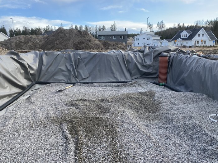 Grävd grop med grusbelagt fundament för pool, omgiven av markduk och en röd plastslang synlig vid sidan.