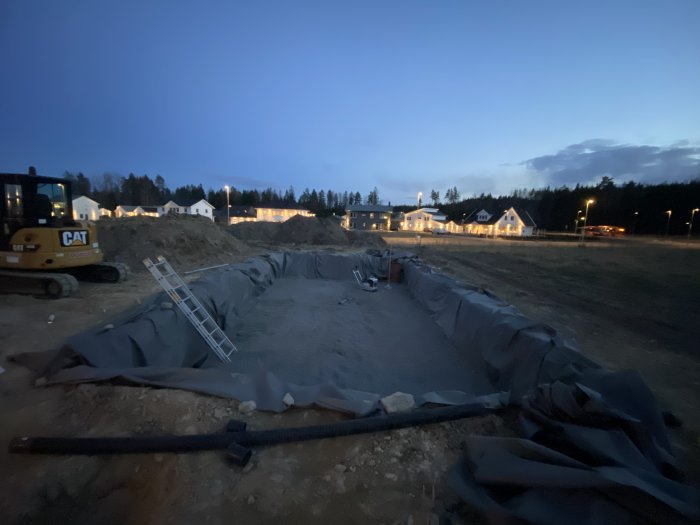 Byggplats vid skymning med grävmaskin och grävt hål i marken förberett för poolinstallation.