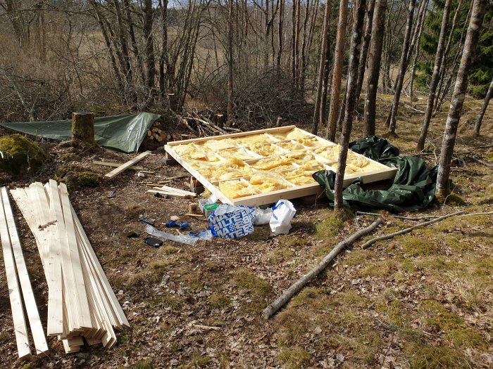 Golvstomme under konstruktion med isolering, omgiven av träplankor och verktyg i skogsmiljö.