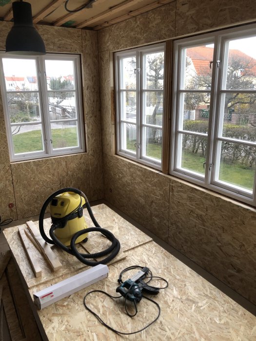 Veranda under renovering med OSB-paneler, fönster, hängande lampa, verktyg och en dammsugare på golvet.