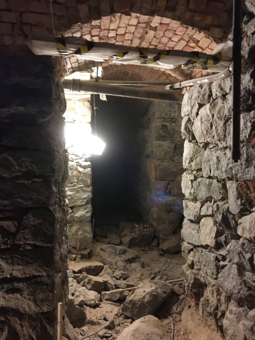 En grävd källargrund med borttaget golv, synliga stenar och byggmaterial, i upplyst arbetsbelysning.