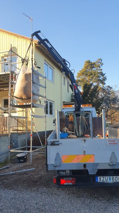 Kranbil lyfter en stor säck byggmaterial framför tvåvåningshus under renovering.