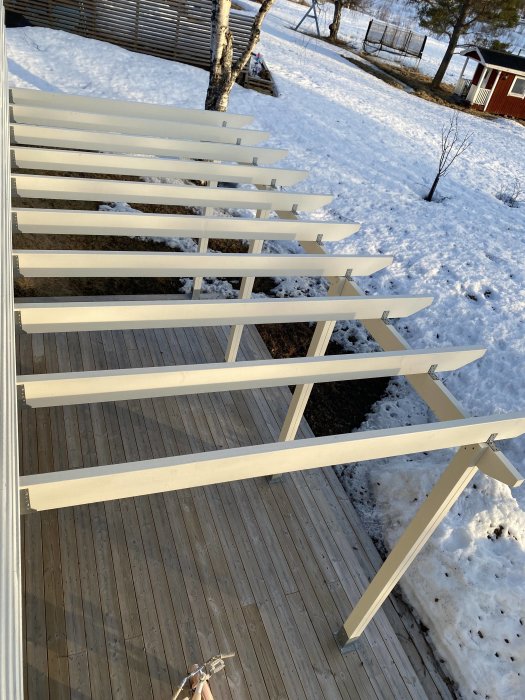 Byggprojekt av en pergola med horisontella vita bräder på en terrass, snö synlig i bakgrunden.