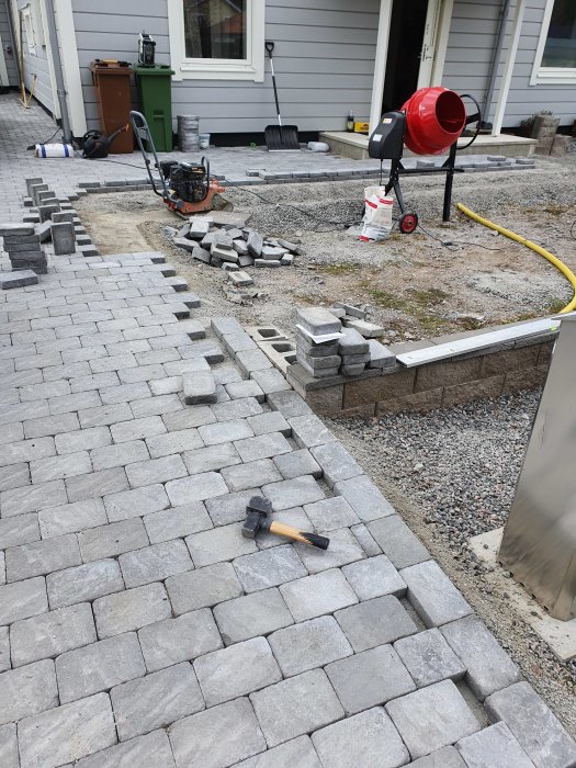 Pågående stenläggning av grå trottoarsten med hammar, cementblandare och kompaktor.