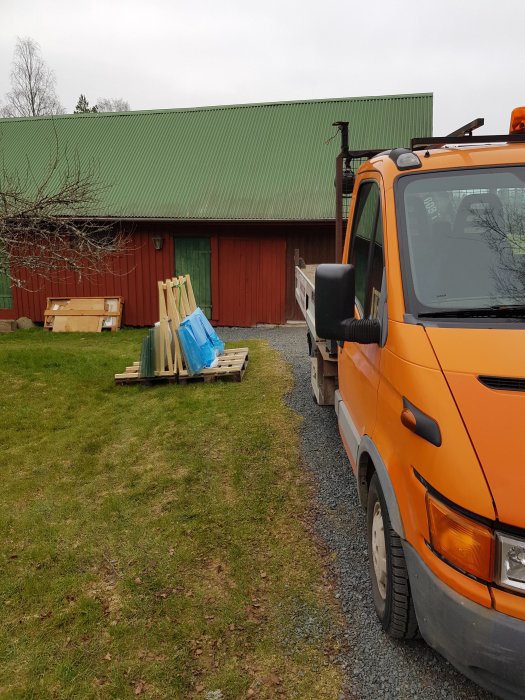 Delar av ett demonterat växthus lastat på pallar bredvid en orange arbetsbil med kran, parkerad på en grusväg.