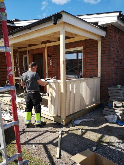 Person bygger en ommålad träfarstukvist vid ett tegelhus under solig dag.