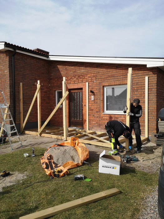 Byggande av en farstukvist i trä framför ett tegelhus, arbetsmaterial och verktyg synliga på gräsmattan.