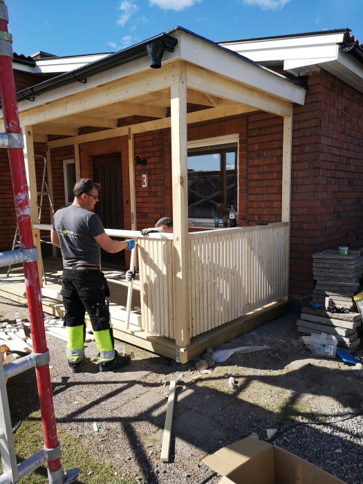 Två personer bygger en träaltan till en tegelvilla under en solig dag.