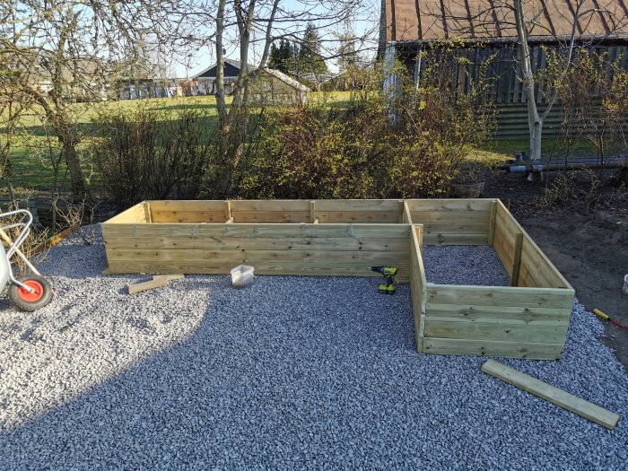 En nybyggd odlingslåda i trä på en grusad yta i en trädgård med verktyg och byggmaterial runtomkring.
