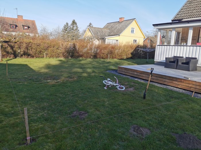 Trädgård med uppmärkt område för odlingshörna, gräsmatta, barnets cykel och trädäck vid hus.