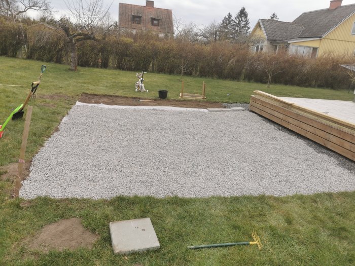 En nyligen grävd yta för odlingshörna och större trädäck med grus, spade, och kratta.