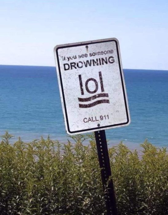 Vägmärke med texten "IF YOU SEE SOMEONE DROWNING LOL" och "CALL 911", havsutsikt i bakgrunden.