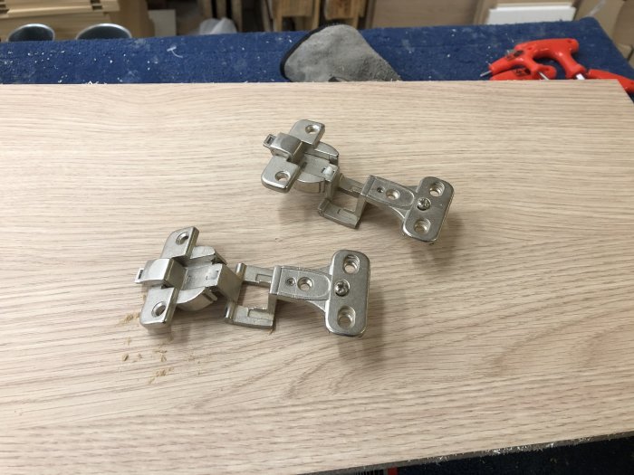 Tre metallgångjärn på en ljus träyta i en verkstadsmiljö, med en röd tång i bakgrunden.