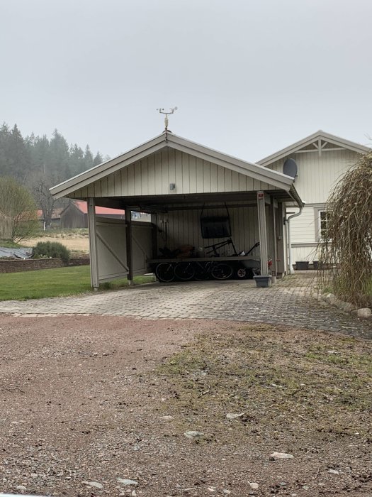 En öppen carport i vit plåt med plats för fordon, angränsande till ett förråd, under en gråmulen himmel.