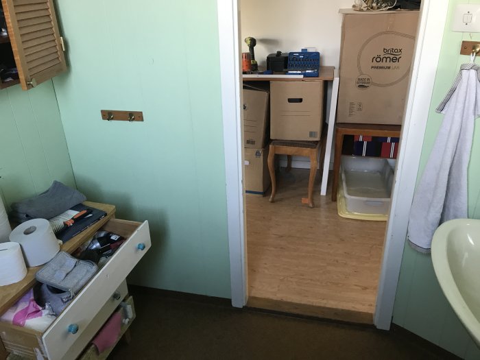 Oorganiserat utrymme med öppen byrålåda, toalettartiklar, flyttkartonger och möbler, som behöver renovering.