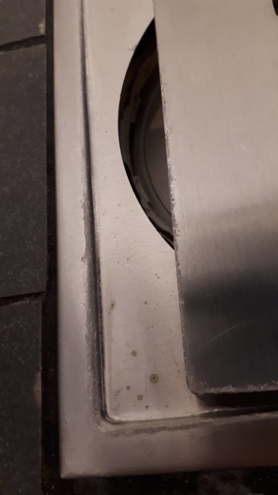 Rostfritt stål golvbrunnssil med vit beläggning på kanterna mot kaklat golv.