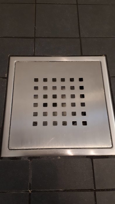 Rostfri golvbrunnssil med vit beläggning på kanter, inbäddad i svart kakelgolv.