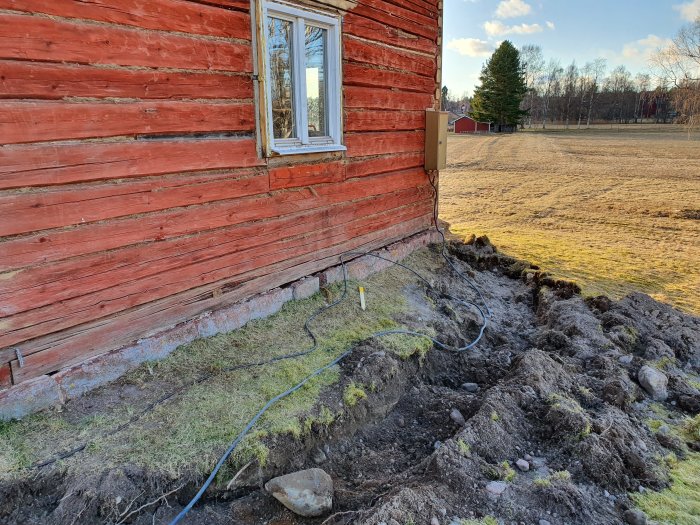 Uppgrävd mark vid röd trävägg där elkabel är omlokaliseras, med utsikt över en äng.