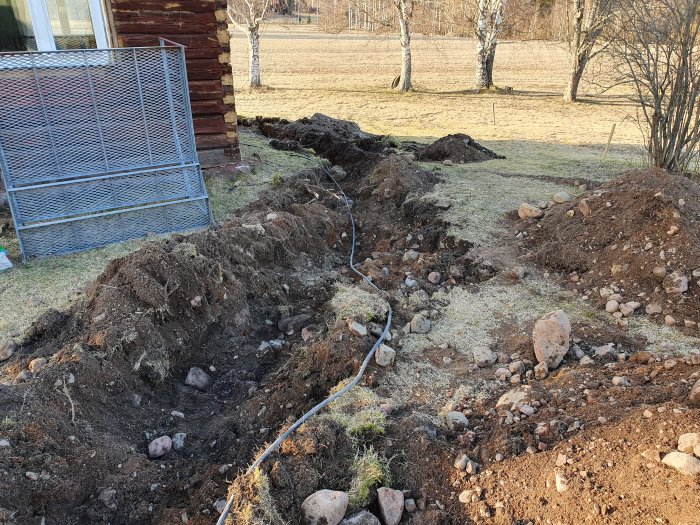 Uppgrävd trädgård med synlig elkabel, stenar och jordhögar nära hus med carport.