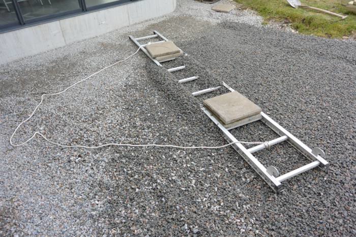 En metallställning med två betongplattor ligger på grus nära en byggnad.
