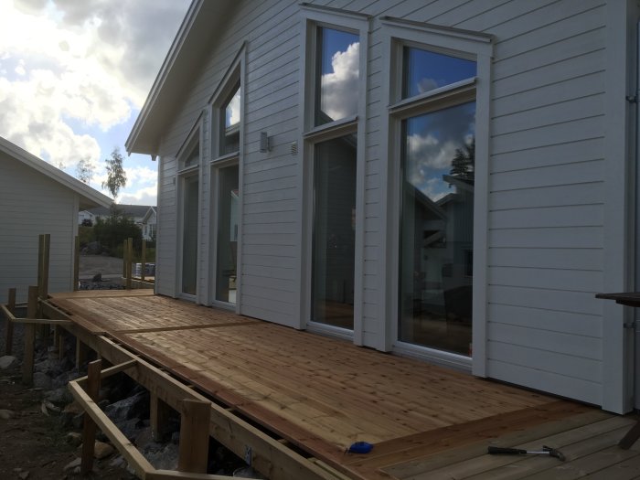 Nybyggd träterrass med stora golvfönster på vit husfasad under klar himmel.