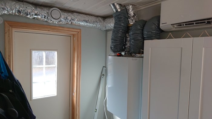 Installation av ventilationssystem med isolerade flexkanaler ovanför dörr och nära luftvärmepump.