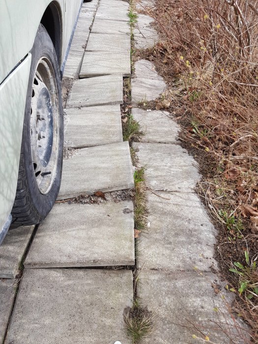 Bild på uppfart med lösa och ojämna betongplattor bredvid bil och visset gräs.