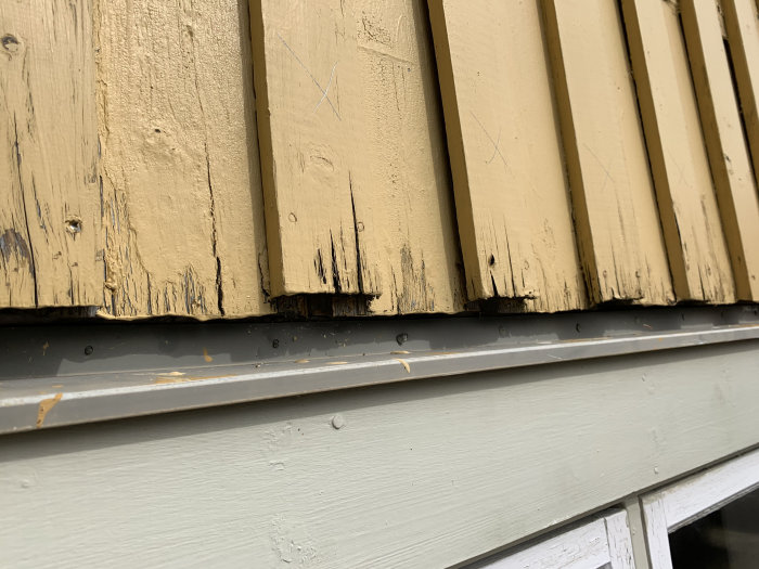 Närbild på en gulmålad träpanel med torrsprickor och slitage vid ändarna ovanför en fönsterbleck.
