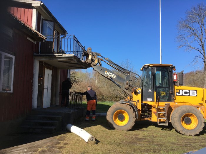 Grävmaskin framför slitet hus med personer som arbetar på rivning av balkong.