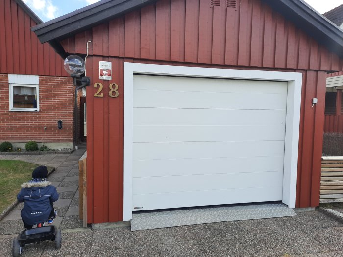 Fasadsiffror på en trästolpe vid ett vitt garage, hus i bakgrunden, barnvagn framför garaget.