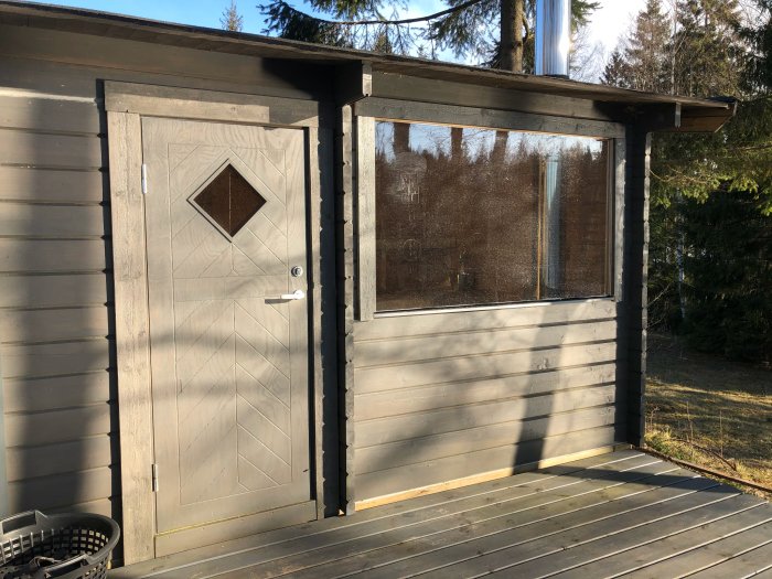 Stort installerat fönster i en grå träfasad bredvid en ytterdörr på en veranda.