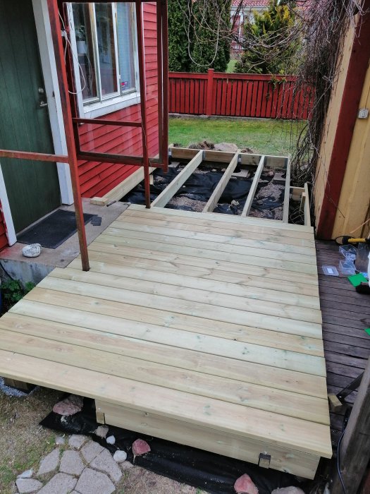 Delvis färdigställd trätrall med terassfötter bredvid ett rött hus och grön trädgård, verktyg synliga.