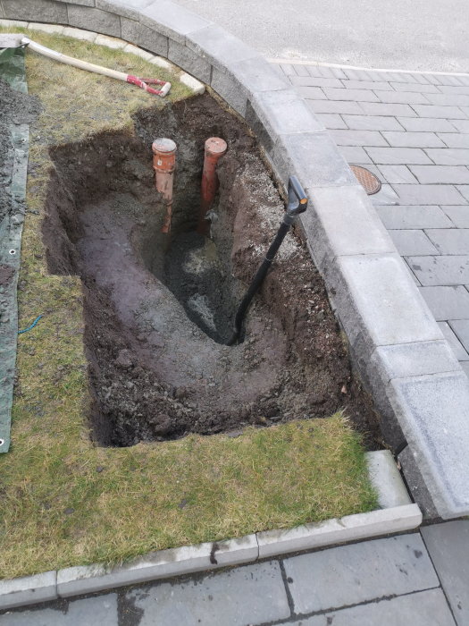 Grävd grop intill en stenmur och trottoarkant, med två synliga rör och verktyg, i sökande efter underjordiska ledningar.