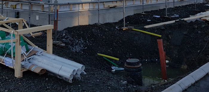 Byggplats med skarvgrop, gula och gröna tomrör, stenmur samt hus i bakgrunden.