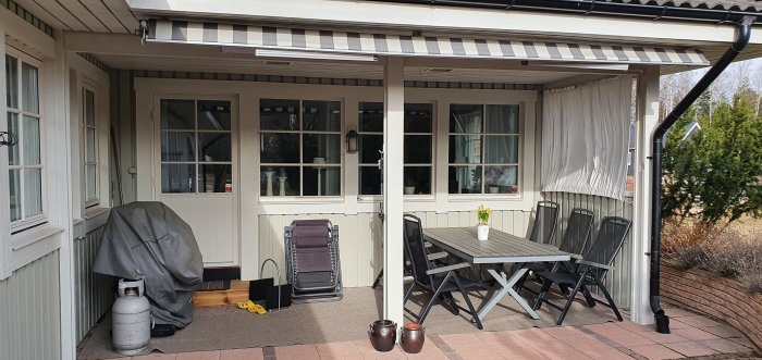 Uteplats med takstöd, befintliga stolpar synliga, och en trämatbord med stolar under markistak nära husvägg.