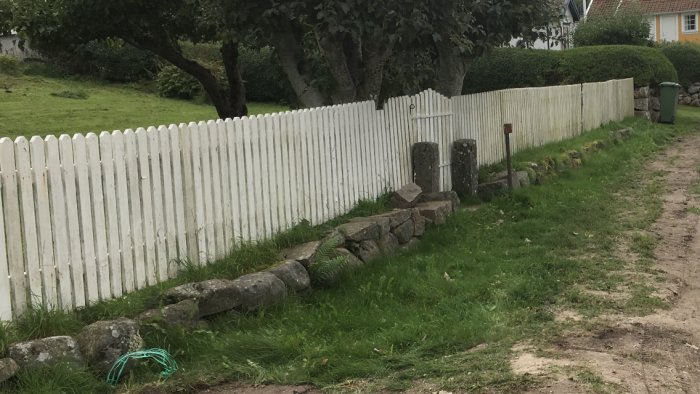 Vitt staket med synliga skador och rost, längs en grusväg med stenmur i förgrunden.