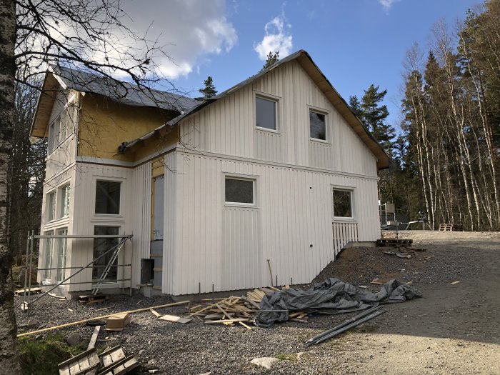 Hus under renovering med partiellt färdigställd vit panel, byggmaterial på marken och omgiven av träd.