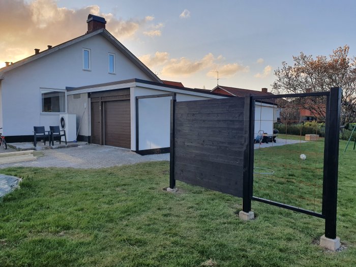 Nymålat mörkt plank framför ett hus, med grön gräsmatta och klar himmel i bakgrunden.