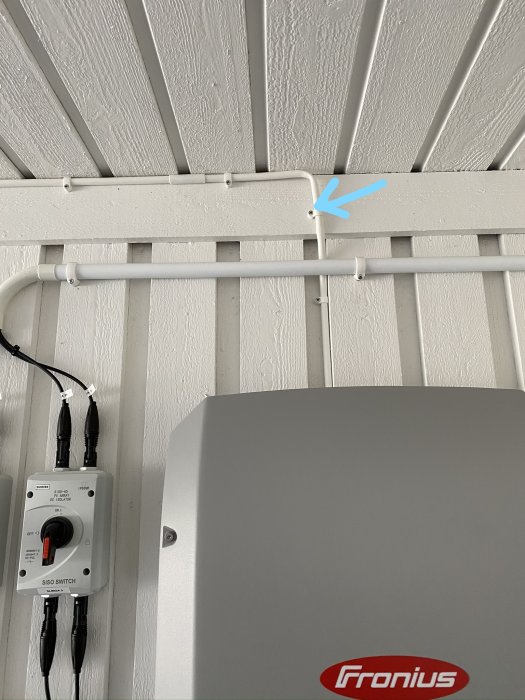 Elinstallation med vit plaströr på vägg, svåråtkomlig kabel markerad med blå pil, nära Fronius-omriktare.