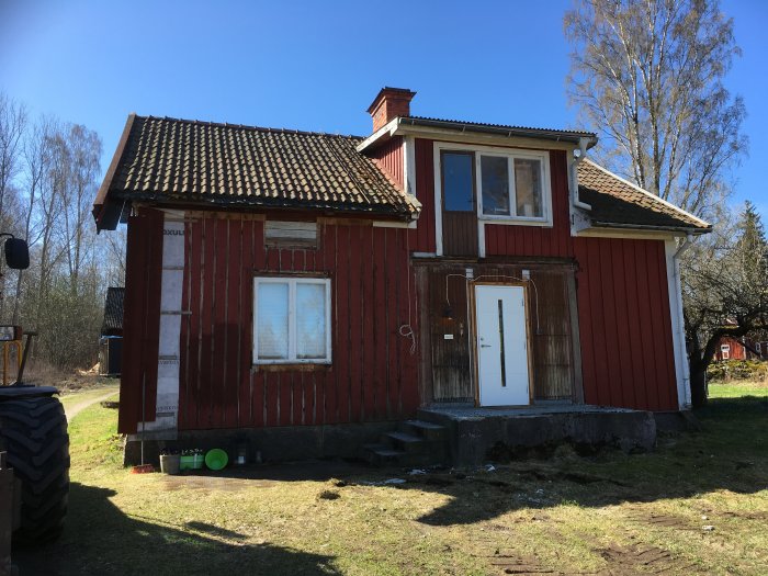 Rött trähus med vitknuten fasad och svart tak; synligt slitage och en delvis renoverad vägg.