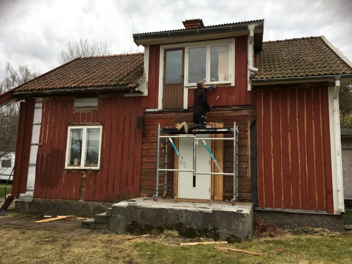Person på byggställning framför röd trähusfasad med synliga renoveringsarbeten och en stenfylld betongklump.