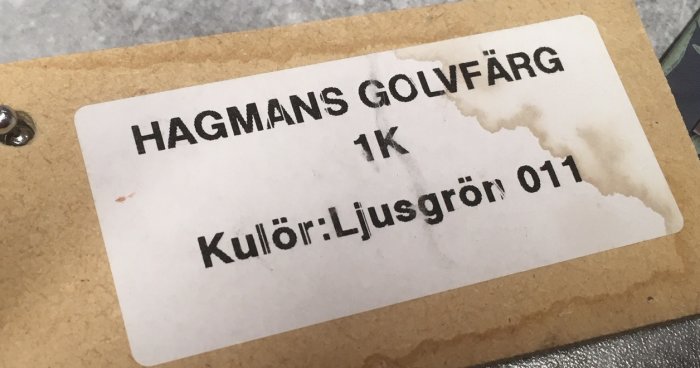 Etikett med texten "HAGMANS GOLVFÄRG 1K, Kulör: Ljusgrön 011" med vattenskada.