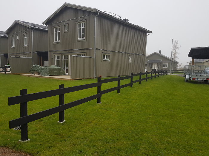 Svart trästaket med horisontella plank och terassering framför grå hus och grönt gräs.