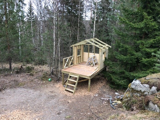 Halvfärdig koja under konstruktion i en skog med plankor och stödstrukturer, utan takbeläggning än.