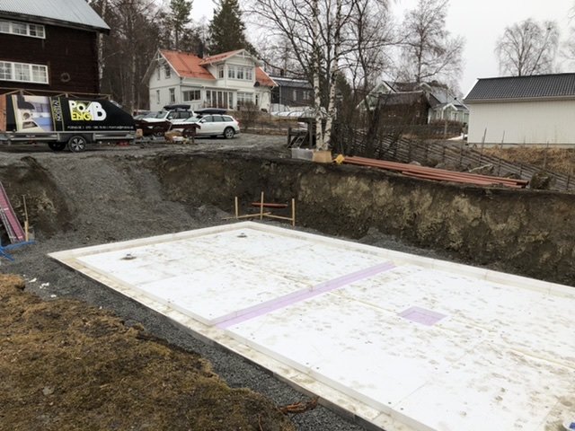 Frigolitisolering och kantelement installerade på en grävd byggplats, med hus och arbetsbil i bakgrunden.