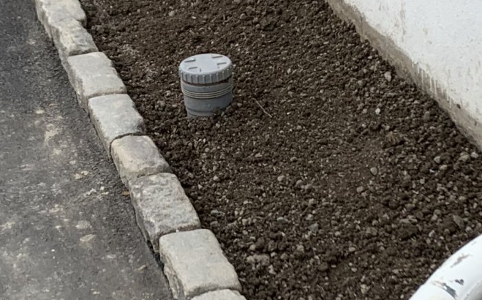 Dräneringslock i mark vid en nyplanterad blomsterrabatt längs en stenlagd kant och vit vägg.